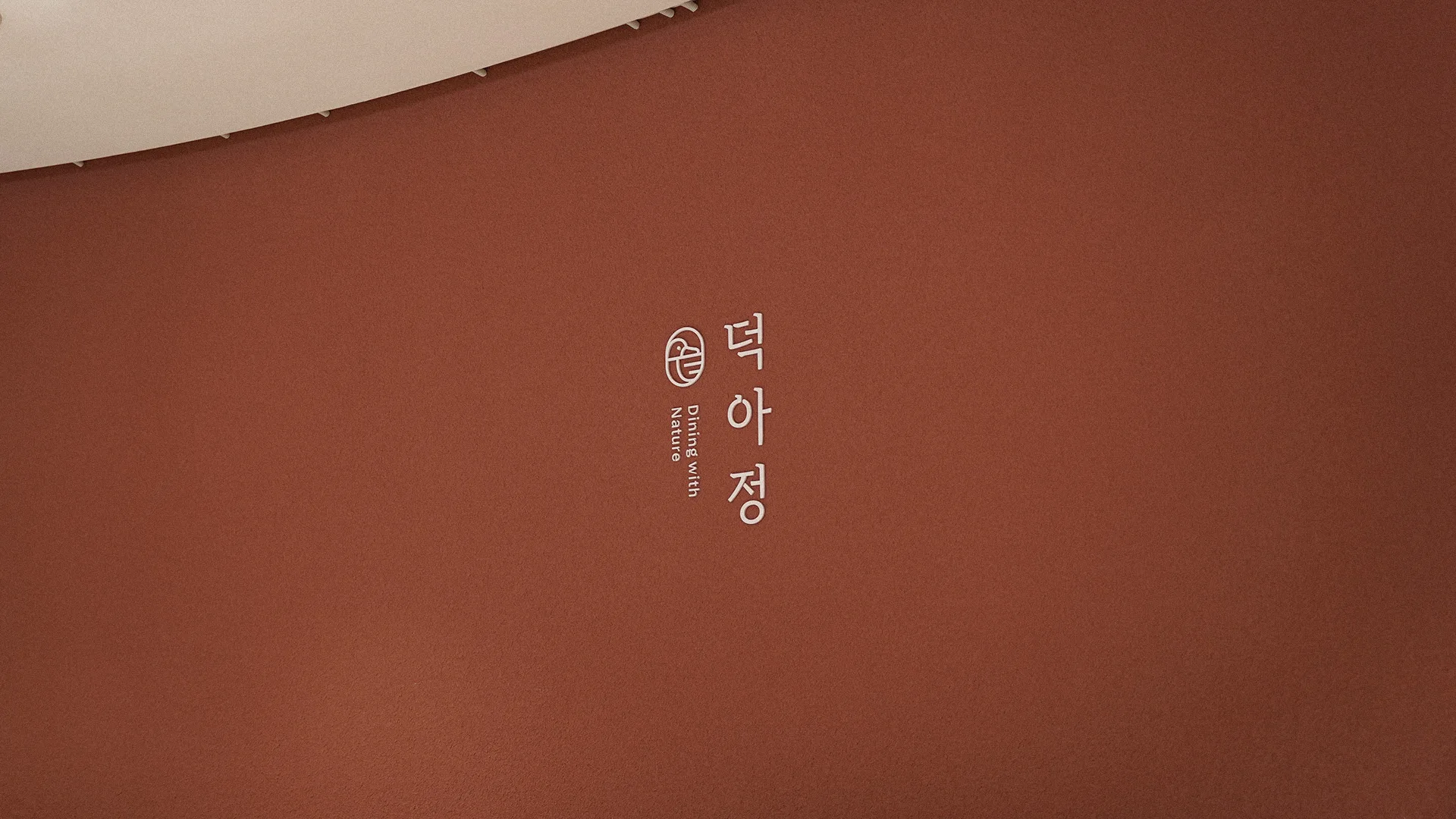 덕아정의 미니멀하고 자연 친화적인 브랜드 아이덴티티가 짙은 갈색 벽면에 흰색 로고와 문구로 표현된 모습입니다. 전통과 현대의 조화를 보여줍니다. 짙은 갈색 벽면에 흰색 덕아정 로고와 'Dining with Nature' 문구가 세로로 새겨져 있습니다.