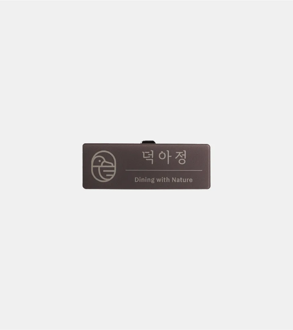 덕아정의 미니멀한 오리 로고와 슬로건이 적용된 명찰 디자인으로, 브랜드의 세련되고 자연 친화적인 이미지를 전달합니다. 덕아정 오리 로고와 'Dining with Nature' 문구가 새겨진 어두운 갈색 명찰.