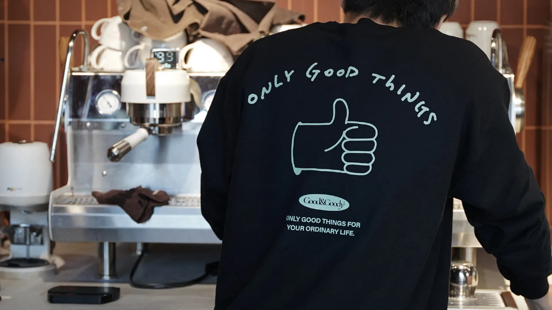 Good&Goody의 슬로건 'ONLY GOOD THINGS FOR YOUR ORDINARY LIFE.'가 새겨진 맨투맨을 입고 카페에서 일상을 즐기는 모습. 브랜드의 친근하고 따뜻한 감성을 담았습니다. 카페에서 'ONLY GOOD THINGS' 문구가 새겨진 검은색 맨투맨을 입은 사람의 뒷모습
