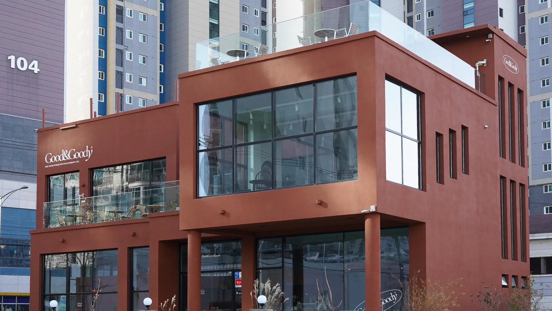 굳앤구디의 따뜻하고 세련된 브랜드 아이덴티티가 반영된 매장 외관. 도심 속 편안한 쉼터의 모습을 보여줍니다. 붉은색 외벽의 굳앤구디 카페 건물, 큰 유리창과 발코니, 흰색 로고.