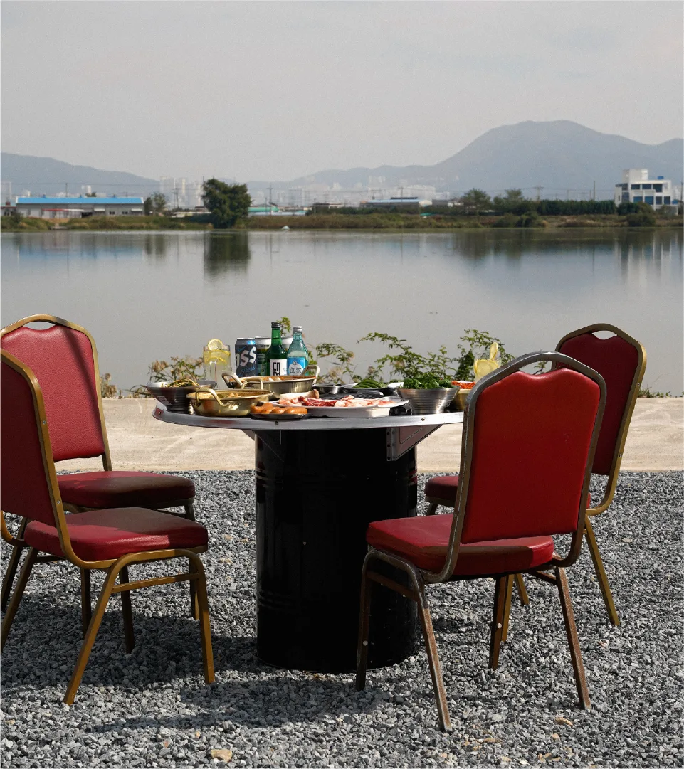 Enjoy Sammi Teukdon in nature. An outdoor dining space by the lake offers a special gourmet experience. Outdoor dining table with grilled pork and drinks by a lake, with red chairs.