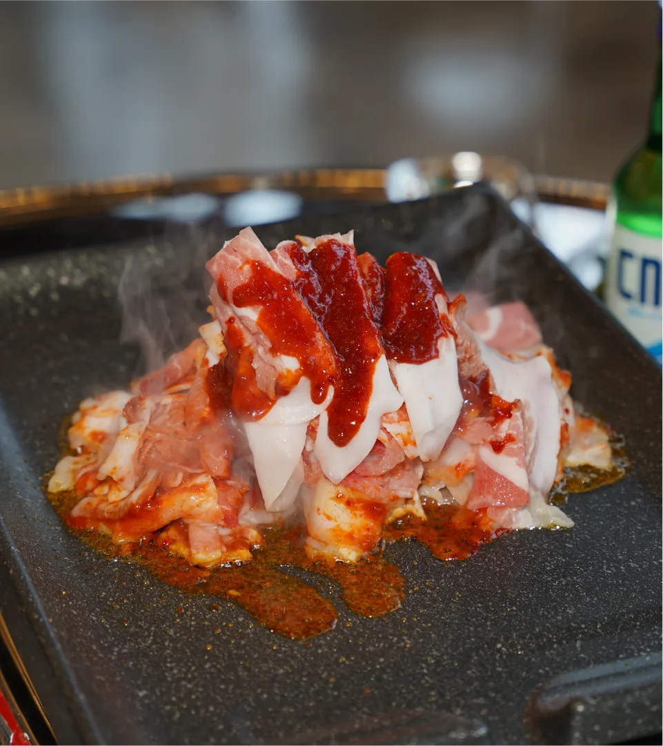 Sammi-Teukdon's rare pork, meeting spicy sauce on the grill, offering a unique gastronomic experience. Thinly sliced pork with spicy sauce grilling on a hot plate, steam rising.
