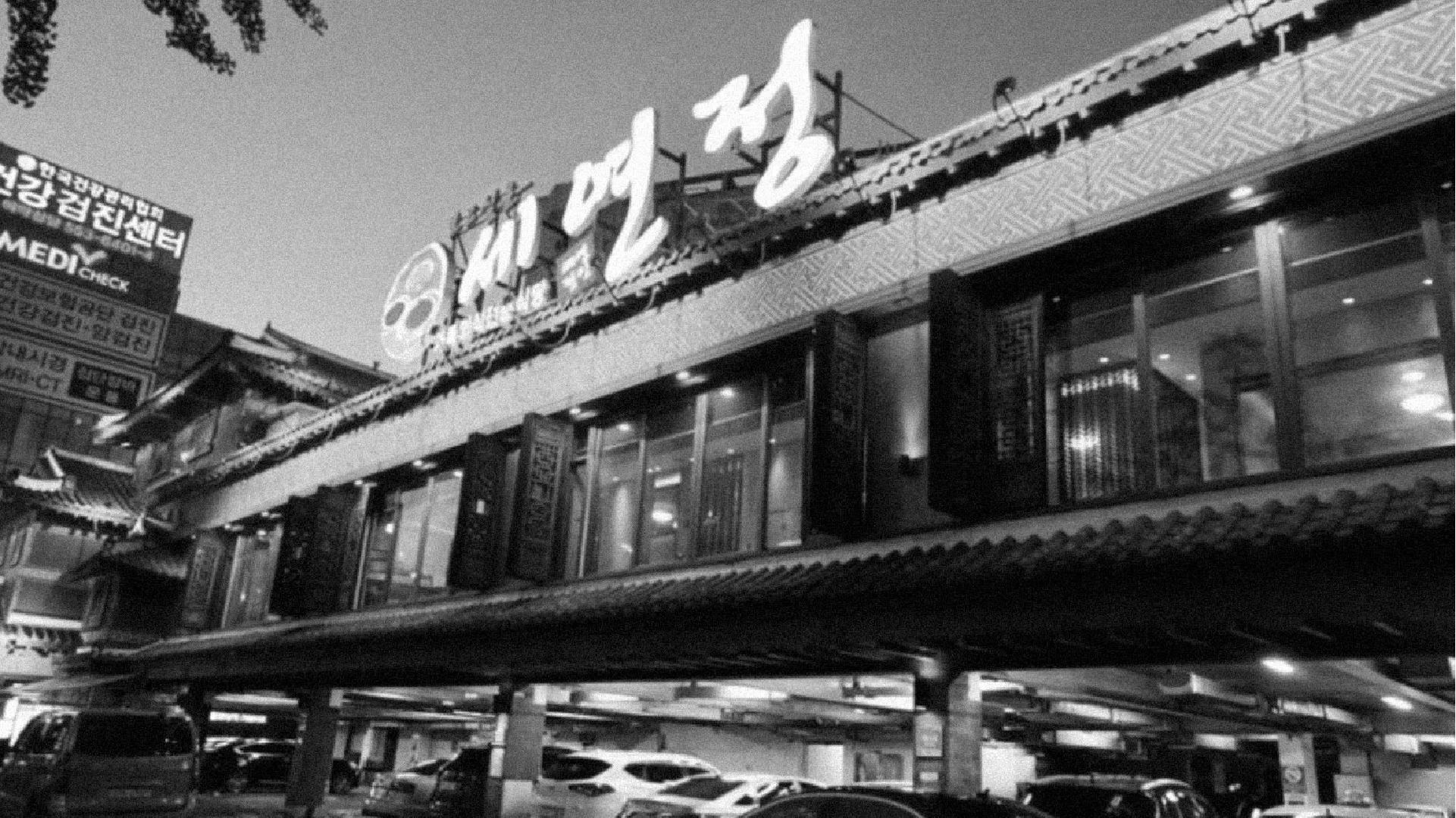 Night view of the 'Seyeonjeong' building, where tradition and modernity coexist. Capturing the authenticity of the space offering Seyeon Chuksan's premium culinary experience. Black and white photo of a building with traditional tiled roofs and modern wind