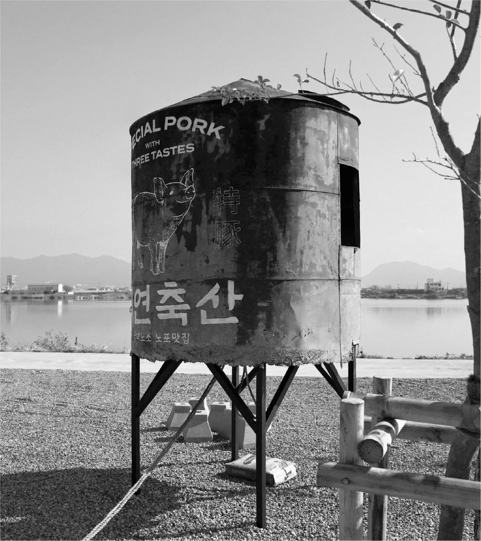 An outdoor sign promoting Seyeon Chuksan's 'Sammi Teukdon'. Its weathered appearance in nature reflects the brand's authenticity and heritage. Black and white photo of a rusty metal pig sign on gravel, with a lake and mount