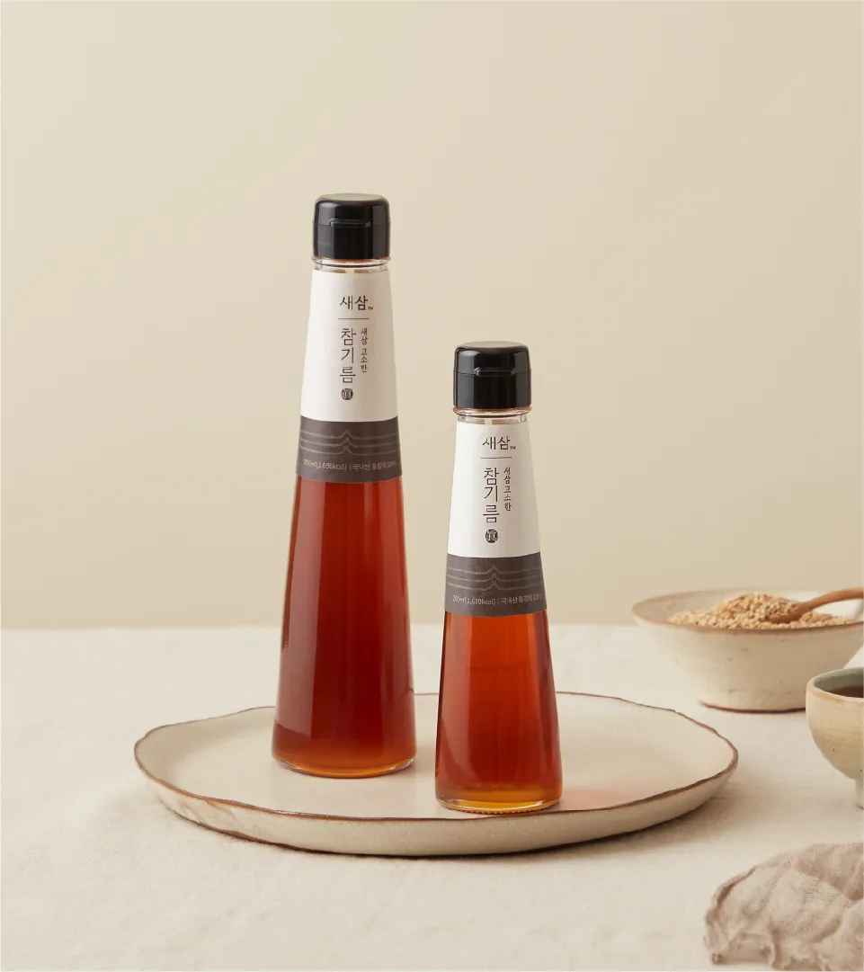 SESAM sesame oil, crafted with 25 years of expertise. Simple and refined design elevates the familiar sesame oil to a premium experience. Two SESAM sesame oil bottles of different sizes on a white plate, with sesame se
